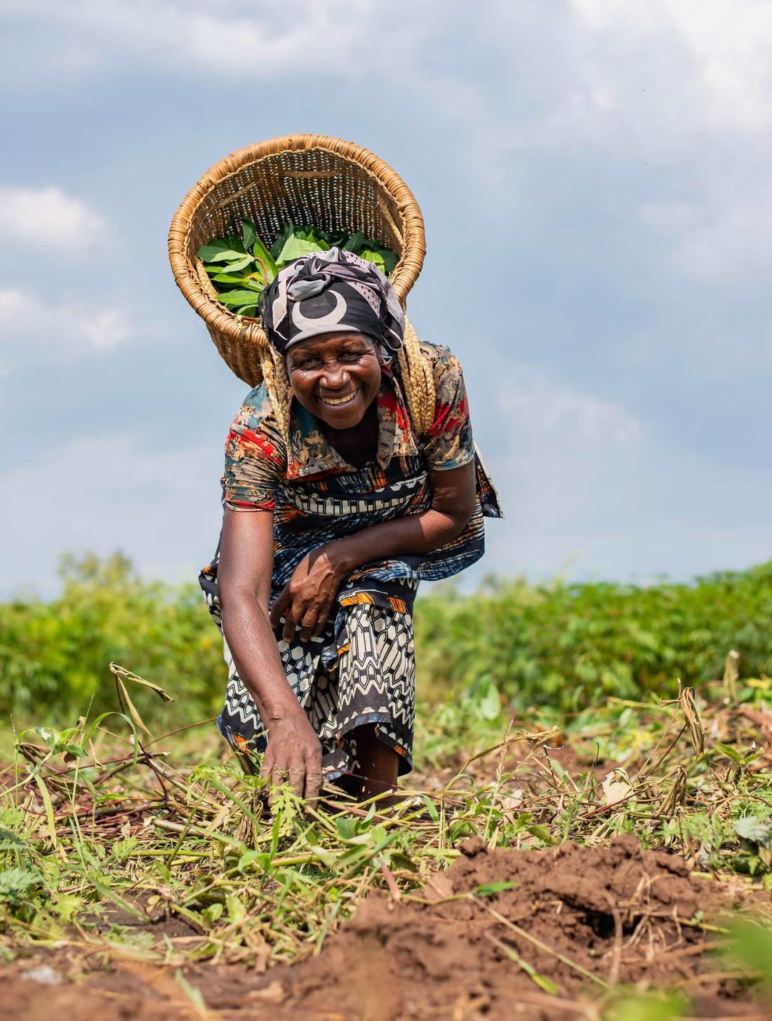 A smiling woman harvesting crops with a woven basket on her back